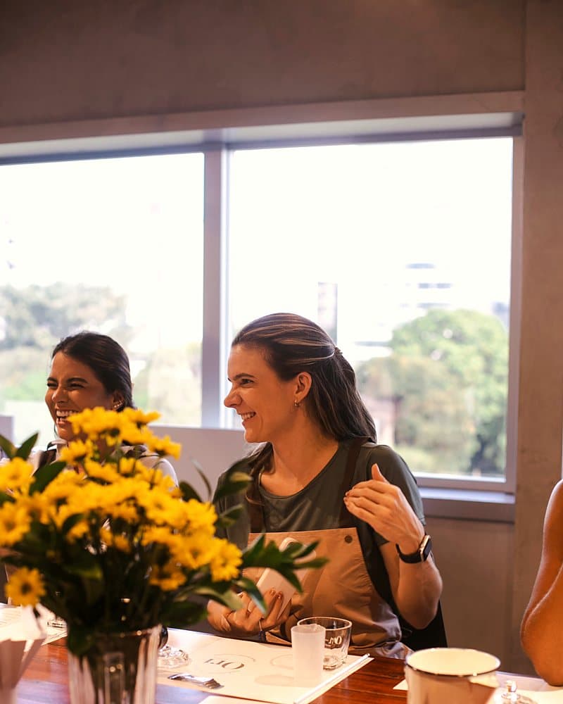 Duas participantes sorrindo na bancada, com as flores do ateliê à frente
